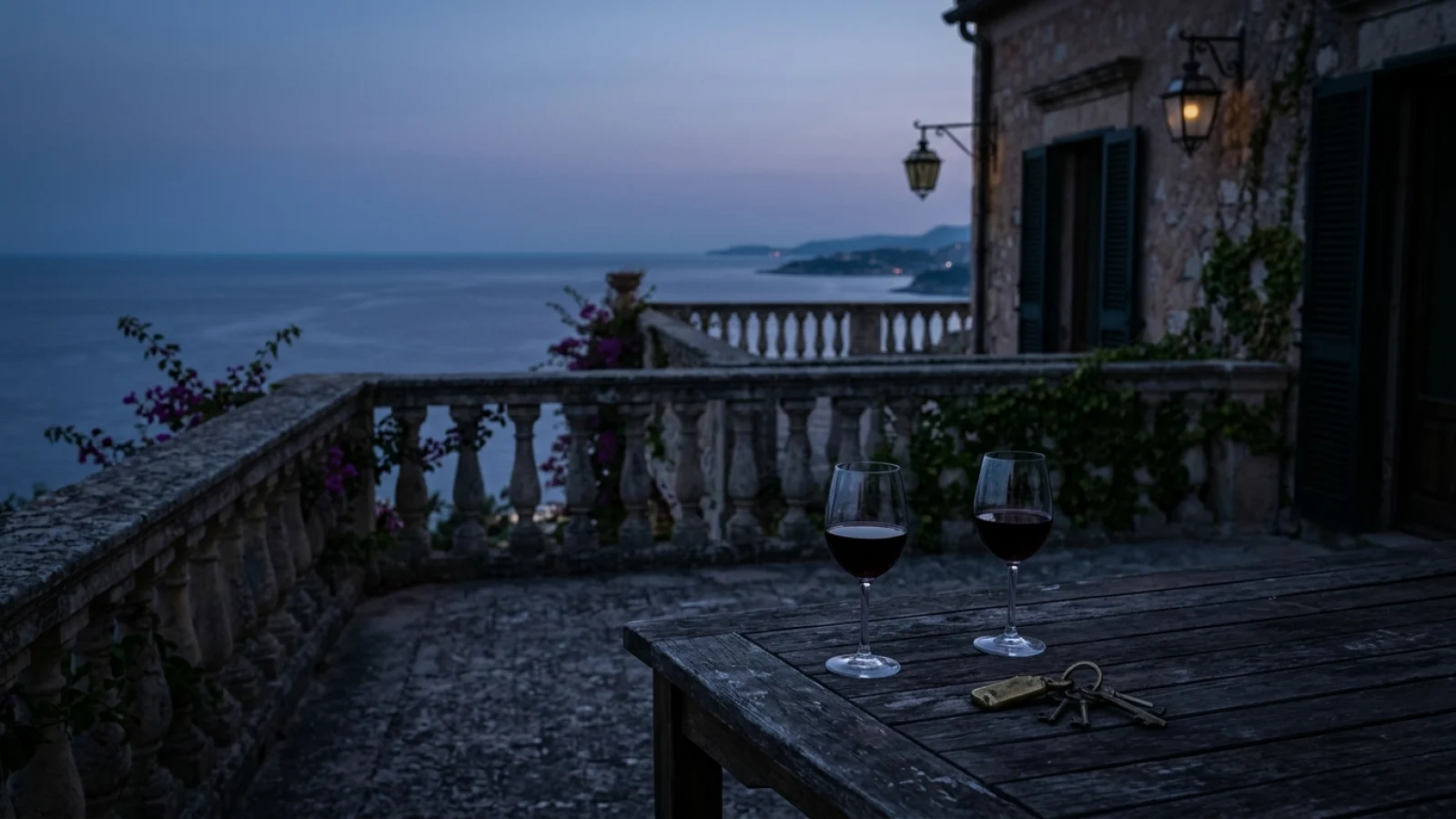 Zwei Weingläser und ein Schlüsselbund auf dem Tisch einer leeren Terrasse mit Meerblick in der Dämmerung.