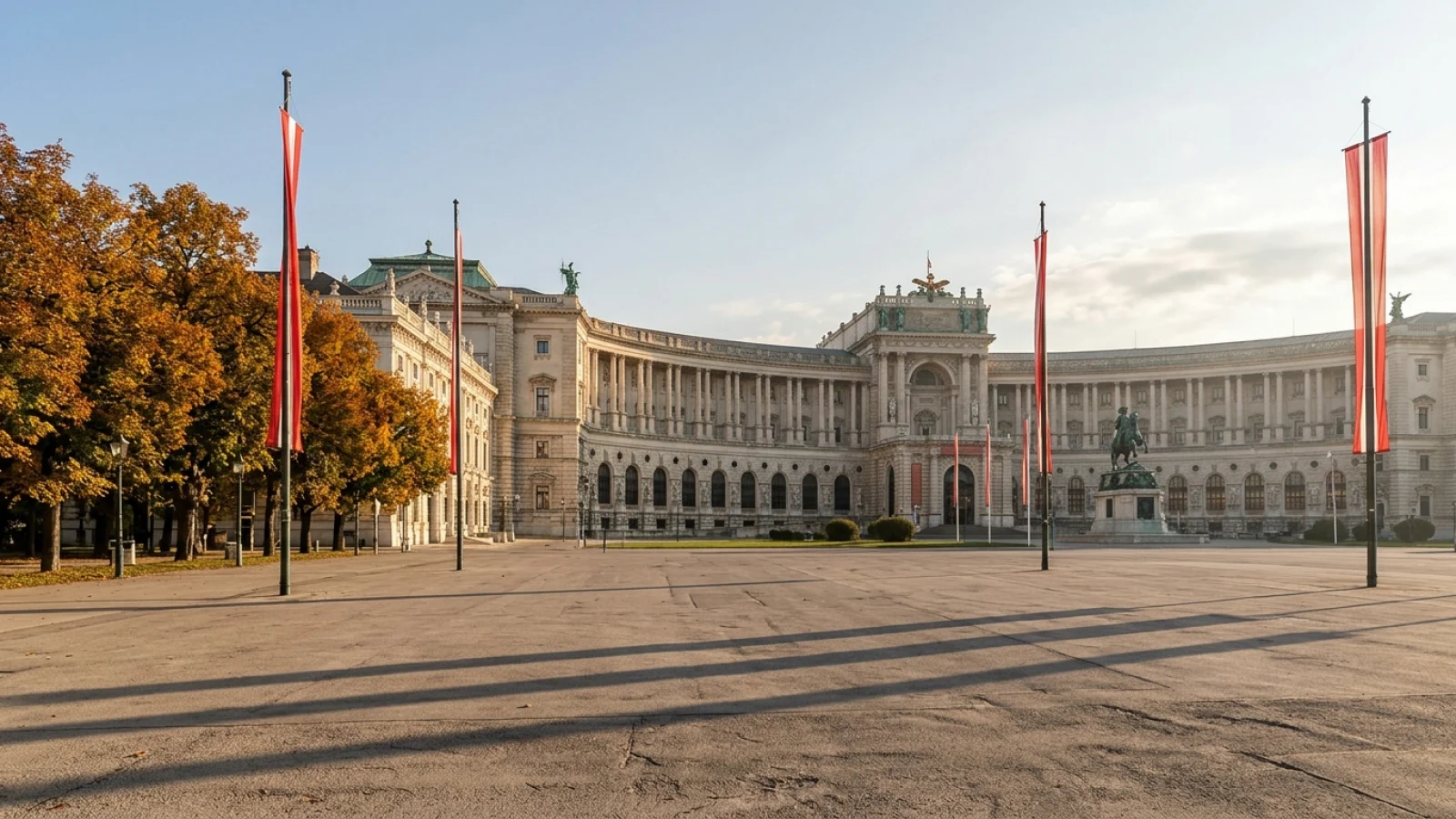 Der leere Heldenplatz in Wien mit der Hofburg im herbstlichen Licht und rot-weiß-roten Fahnen an Masten.