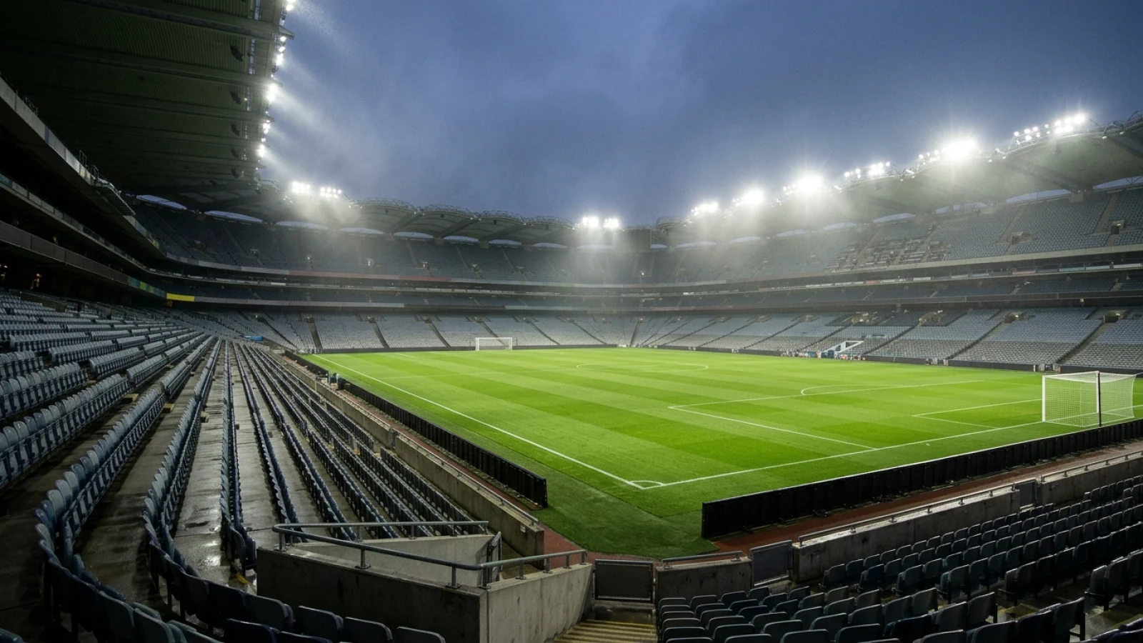 Ein leeres Fußballstadion am Abend unter hellen Flutlichtern mit verwaisten Rängen und grünem Rasen.