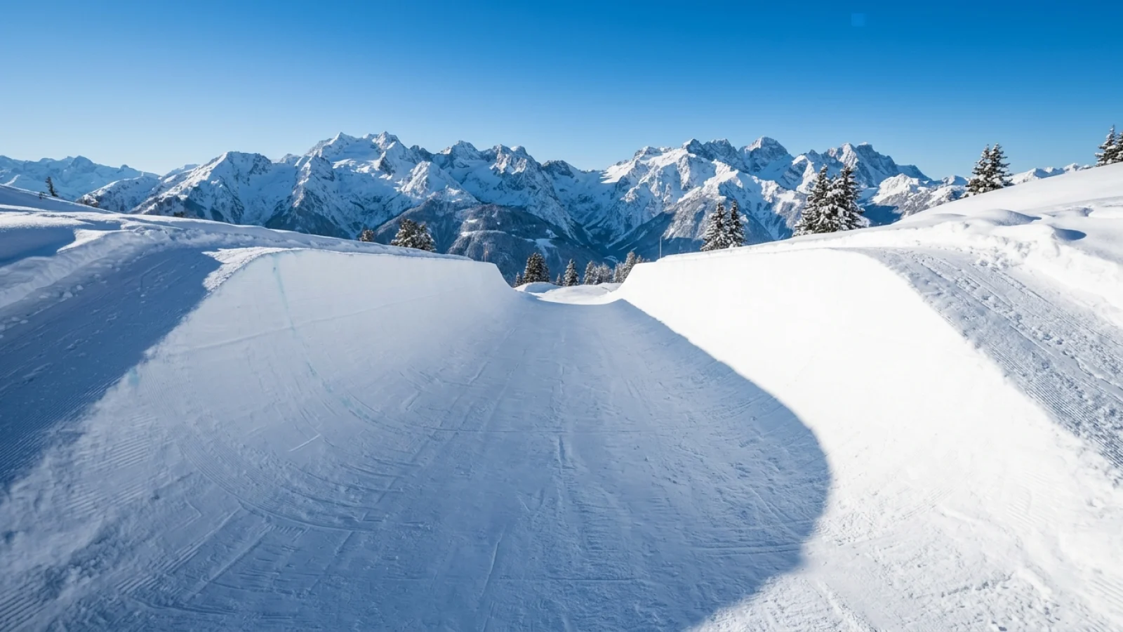 Leere Snowboard-Halfpipe mit glatt präparierten Schneewänden vor einem alpinen Bergpanorama im Sonnenlicht.